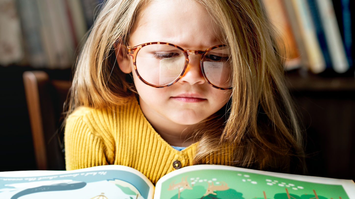 Niña usando anteojos redondos de carey leyendo un libro infantil en biblioteca